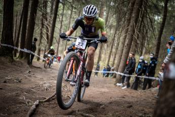 Christopher Blevins wears a tight fitting biking outfit and helment as he rides a mountain bike in a forest. There are tall trees behind him.