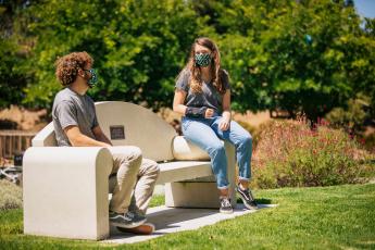 Two students wearing face masks sit on opposite ends of a concrete bench outdoors. 