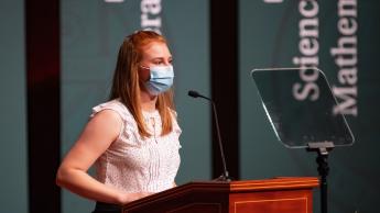 A woman wearing a face covering speaks at a podium in front of a telepromoter 