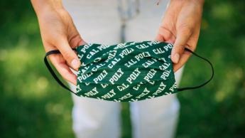 A person just out of frame holds out a green Cal Poly branded face covering