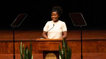 A smiling young woman in a white dress stands behind a podium on a stage