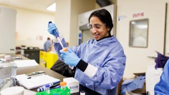 A student in a blue smock and safety glasses uses a pipet to transfer liquid into a flask in a biomedical engineering lab