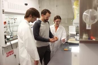 A professor stands between two students in lab coats as they handle debondable glue sticks in a chemistry lab.