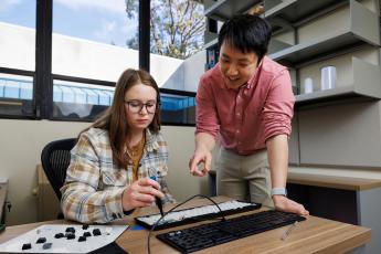 A professor stands with a student who is repairing a keyboard at a desk