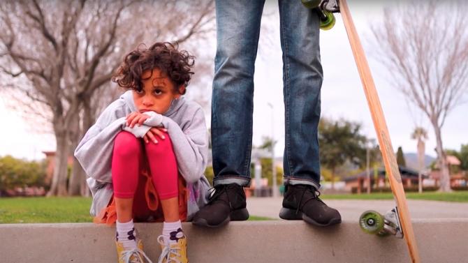 Still image from a student film of a young child sitting on a curb next to a caregiver who is holding a skateboard. 