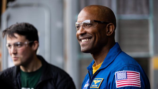 Captain Victor Glover smiles while wearing safety glasses and a blue flight suit in the Cal Poly aerospace engineering hangar